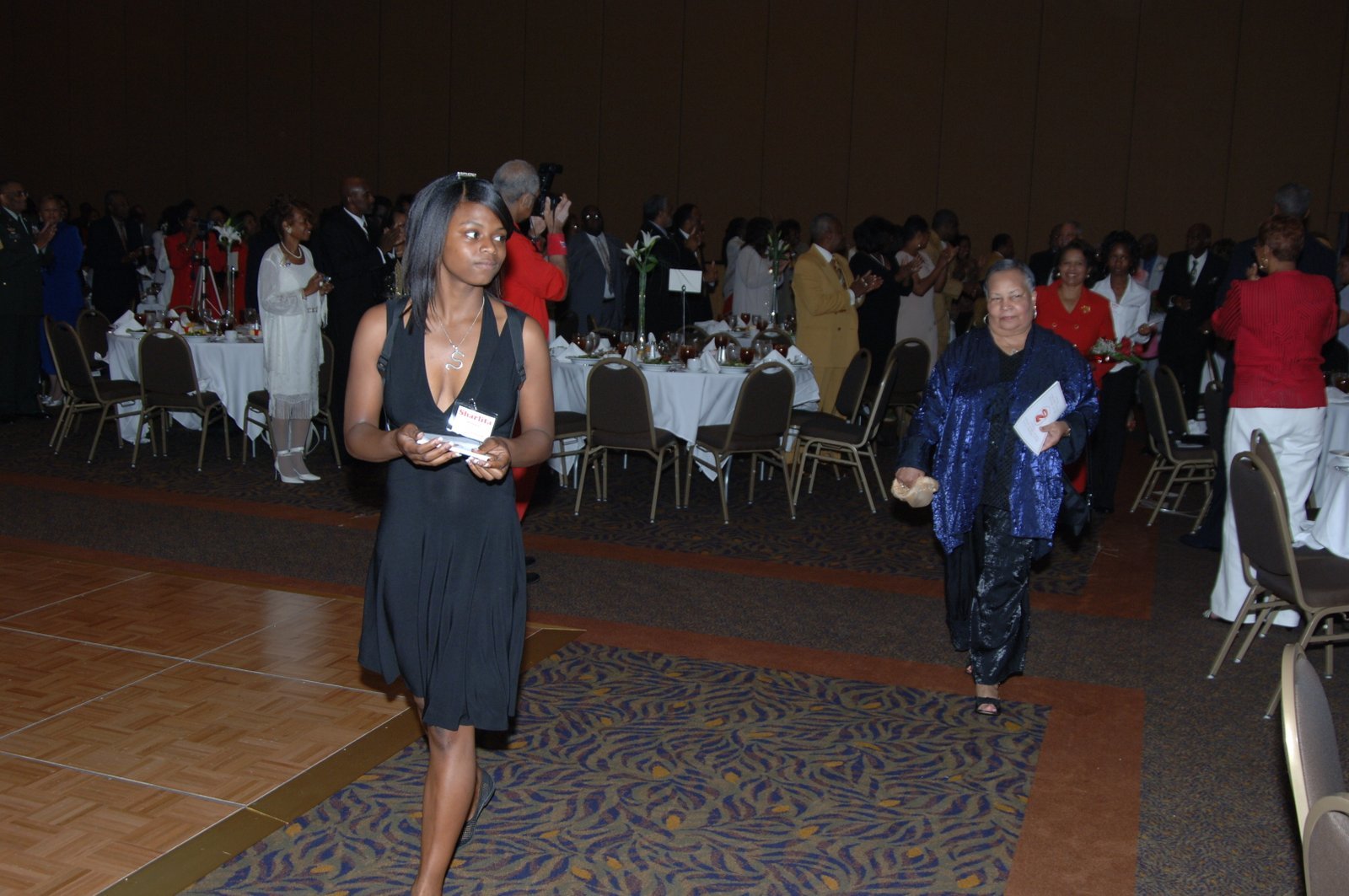 Woman in black dress and woman in blue dress at formal event with tables and chairs